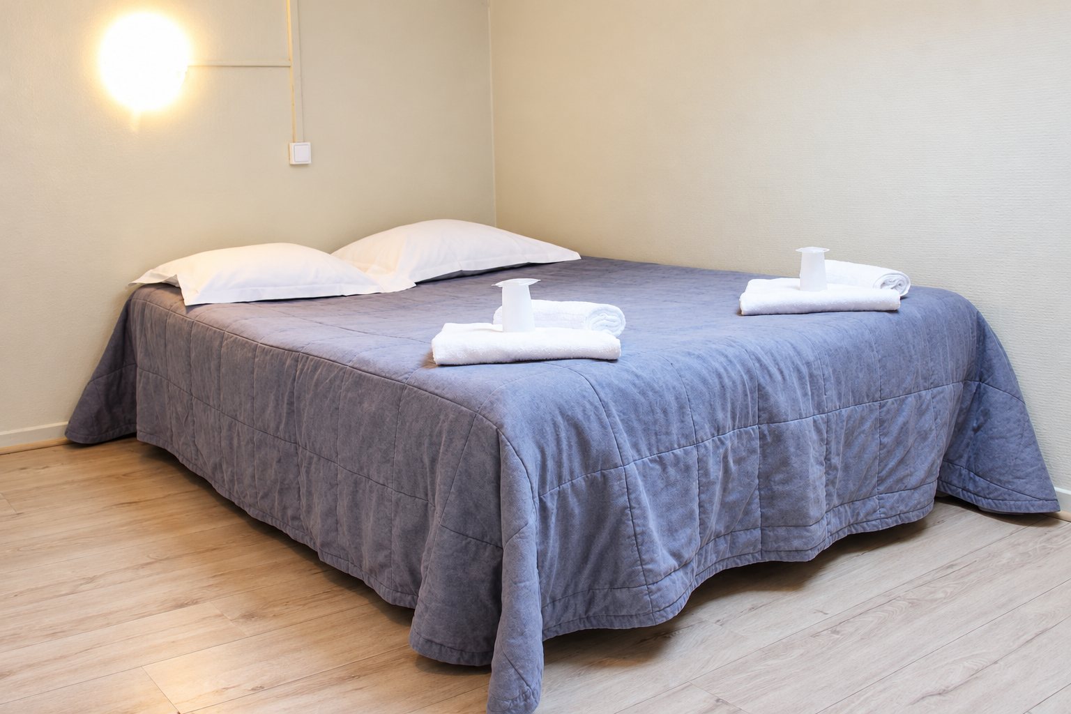 Une chambre épurée avec un grand lit recouvert d'un couvre-lit gris-bleu, deux oreillers blancs et des serviettes blanches pliées sur le dessus.