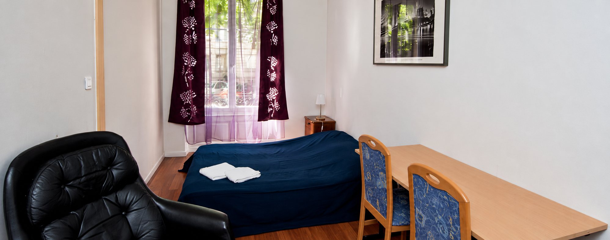 Chambre lumineuse avec un lit recouvert d'un couvre-lit bleu marine, des serviettes blanches, un bureau en bois, deux chaises et un fauteuil en cuir noir devant une fenêtre aux rideaux violets.