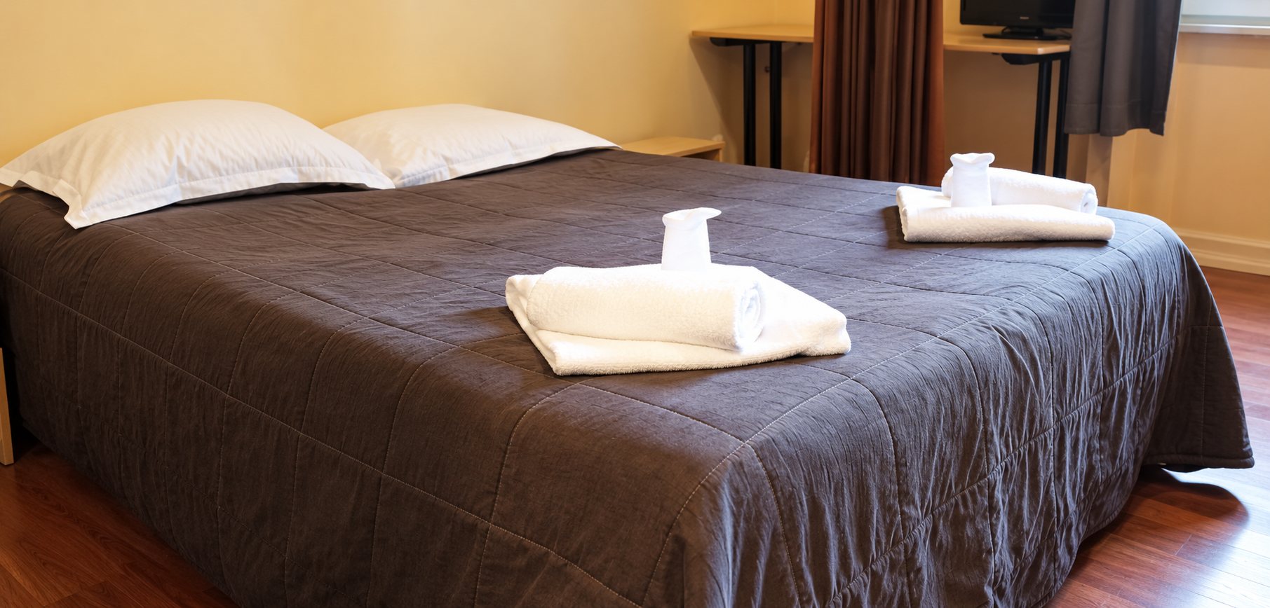 Un lit double recouvert d'un couvre-lit gris foncé avec des serviettes blanches pliées posées dessus, dans une chambre au parquet en bois.