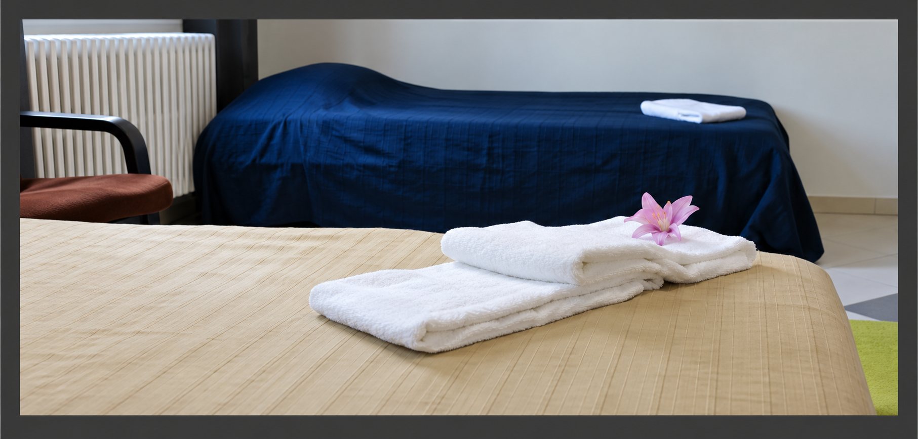 Une chambre avec deux lits dont l'un porte des serviettes blanches pliées décorées d'une fleur rose, près d'un radiateur.