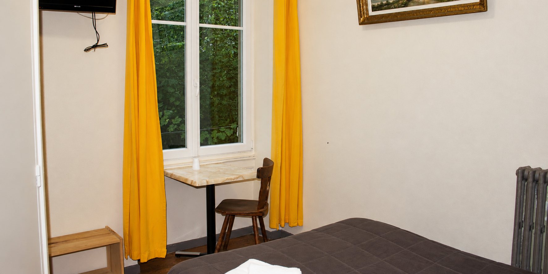 Chambre d'hôtel simple avec un lit couvert d'un plaid marron, une petite table près d'une fenêtre aux rideaux jaunes et un radiateur en fonte.