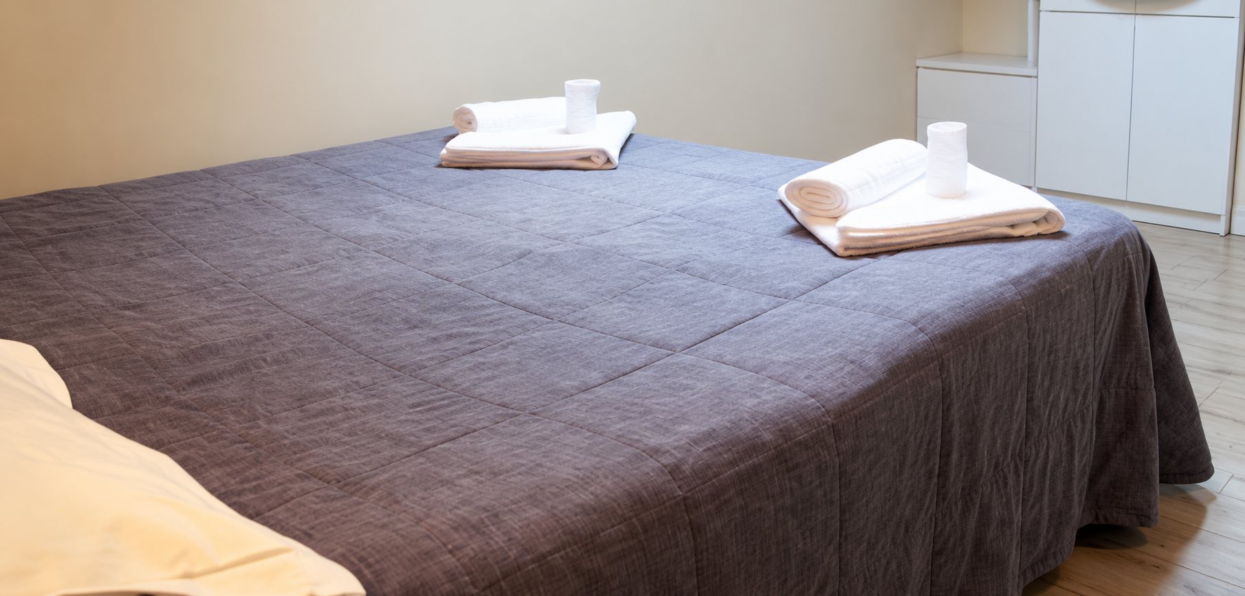 Un grand lit double recouvert d'un couvre-lit gris foncé, avec des serviettes blanches pliées posées dessus, dans une chambre lumineuse.