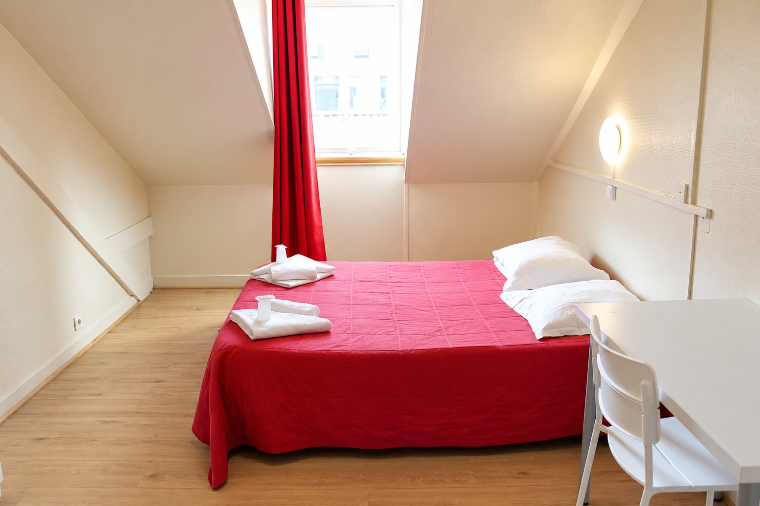 Chambre sous les combles avec un lit double couvert d'un tissu rouge, des serviettes blanches pliées, un rideau rouge, un bureau et une chaise blancs.