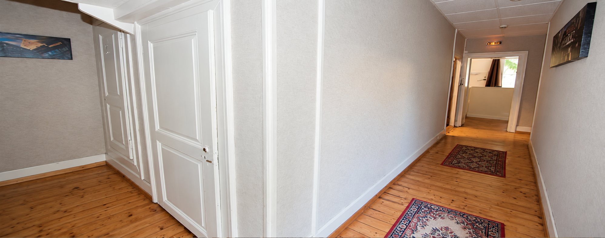 Un long couloir aux murs clairs avec un parquet en bois, deux petits tapis, des portes blanches et un tableau accroché au mur.