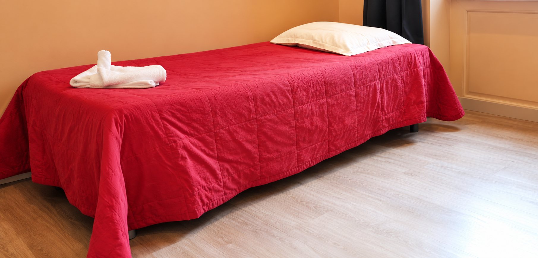 Un lit simple recouvert d'un couvre-lit rouge, avec un oreiller beige et une serviette pliée, posé sur un parquet clair dans une chambre aux murs orangés.