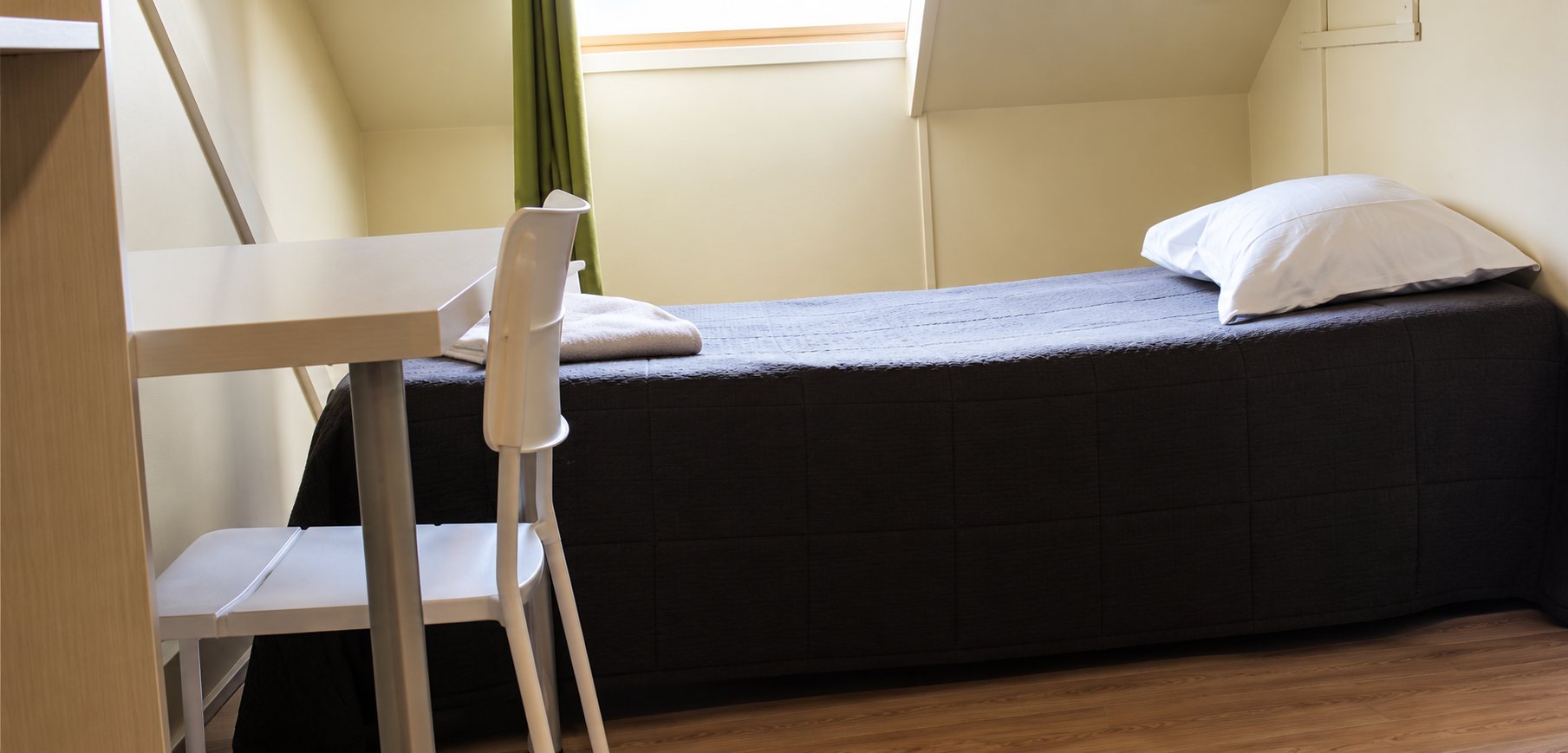 Une petite chambre mansardée avec un lit simple recouvert d'un drap sombre, un oreiller blanc, un bureau et une chaise blancs.